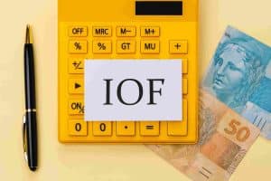 Calculadora amarela com cédulas de real e um cartão com a sigla IOF em destaque, ao lado de uma caneta preta, representando o impacto do IOF nas transações financeiras e sua influência sobre operações econômicas no Brasil.