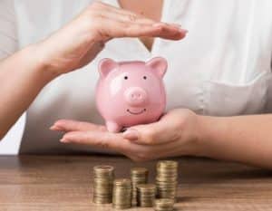 Pessoa segura um cofre em formato de porquinho com as mãos em gesto de proteção, diante de pilhas de moedas sobre a mesa, simbolizando o cuidado necessário para proteger seu patrimônio e manter a segurança financeira.