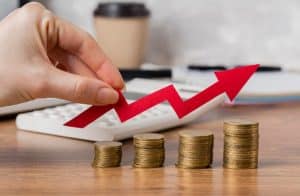 Mão segurando uma seta vermelha apontando para cima sobre pilhas crescentes de moedas, simbolizando o aumento de preços e seus efeitos na economia e no poder de compra da população.