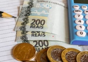 Notas de duzentos reais, moedas de um real e uma calculadora azul dispostos sobre um caderno com linhas, ao lado de um lápis apontado. A composição remete ao controle financeiro cotidiano, com destaque para o cálculo de despesas e planejamento orçamentário. A imagem simboliza a importância de organização ao lidar com compras parceladas, ressaltando o papel da análise consciente para evitar dívidas e manter as finanças sob controle.