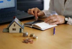 Pessoa analisa finanças pessoais utilizando calculadora e caderno, com moedas empilhadas e uma maquete de casa sobre a mesa, representando o controle da economia doméstica e o planejamento financeiro familiar.