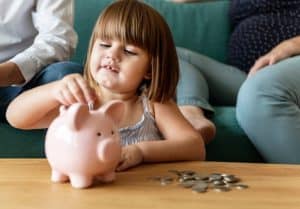 Criança sorridente insere uma moeda em um cofrinho rosa, sentada à mesa com várias moedas espalhadas ao redor, enquanto adultos observam ao fundo. A cena transmite um momento de aprendizado e incentivo à construção de hábitos financeiros desde cedo. A imagem representa o valor da educação financeira infantil, destacando a importância de ensinar, de forma lúdica e prática, conceitos como economia, responsabilidade e planejamento.