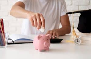 Pessoa deposita moeda em cofrinho rosa sobre a mesa, ao lado de livros, diploma e materiais escolares, simbolizando o valor da educação financeira no planejamento de metas e no futuro acadêmico.