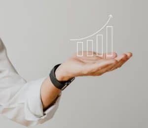 Mão estendida de uma pessoa com camisa social branca e smartwatch sustenta um gráfico de barras crescentes com seta apontando para cima, representando crescimento e progresso. A ilustração flutuante simboliza o conceito de evolução financeira, sugerindo estabilidade, planejamento estratégico e aumento gradual de resultados econômicos.