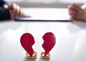 Duas alianças douradas sobre uma mesa sustentam um coração vermelho rasgado ao meio, simbolizando a separação de um casal. Ao fundo, duas pessoas estão sentadas frente a frente, em ambiente formal, sugerindo mediação ou processo legal. A imagem representa o desafio de organizar finanças em caso de divórcio ou separação, incluindo divisão de bens, acordos patrimoniais e reestruturação da vida financeira individual.