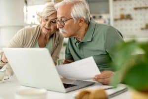 Casal de idosos sentado à mesa observa atentamente documentos enquanto utiliza um notebook. Ambos demonstram concentração e colaboração, representando o planejamento conjunto e o cuidado com as finanças na terceira idade, como controle de gastos, aposentadoria, investimentos e segurança financeira na fase madura da vida.
