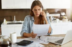 Mulher jovem com expressão preocupada analisa contas e boletos enquanto fala ao telefone, sentada à mesa da cozinha. À sua frente estão papéis, calculadora, caneca, cafeteira e notebook, simbolizando o desafio cotidiano de organizar e equilibrar as finanças pessoais em meio às responsabilidades domésticas.