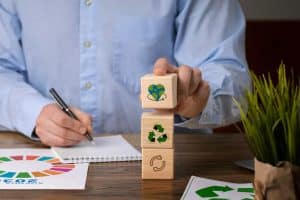 Pessoa com camisa azul organiza blocos de madeira com ícones relacionados à sustentabilidade, como reciclagem, economia circular e um coração envolvendo o planeta Terra. Sobre a mesa há documentos coloridos e um caderno de anotações, simbolizando planejamento e compromisso com práticas de finanças sustentáveis em alinhamento com os Objetivos de Desenvolvimento Sustentável da Agenda 2030.