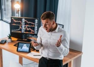 Homem de expressão preocupada e postura exaltada olha para o celular em um escritório, enquanto gráficos de queda são exibidos em três telas ao fundo. A cena sugere perdas financeiras inesperadas, ilustrando o impacto emocional e prático de crises no mercado ou fraudes digitais. A imagem retrata uma possível vítima de golpes financeiros, ressaltando a importância de vigilância, planejamento e segurança na gestão dos investimentos.