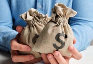 Pessoa segura dois sacos de tecido com símbolo de cifrão, representando a importância de investir e poupar como formas complementares de construir e proteger o patrimônio financeiro ao longo do tempo.