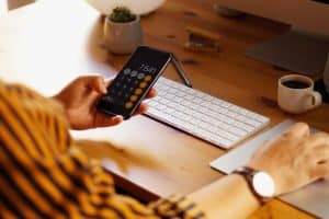 Pessoa utiliza aplicativo de calculadora no celular ao lado de computador e teclado, representando o acompanhamento de finanças pessoais e a tomada de decisões com foco em investimentos de liquidez diária.