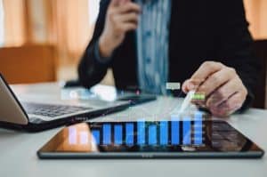 Pessoa em traje formal utiliza tablet e notebook sobre a mesa, com gráficos financeiros e indicadores holográficos projetados sobre a tela do dispositivo. A imagem representa análise de dados e acompanhamento de desempenho econômico. Com forte apelo visual à tecnologia e à tomada de decisões estratégicas, a cena simboliza a atuação no mercado de crédito, onde análises precisas e comportamento dos indicadores são fundamentais para avaliar riscos e oportunidades de investimento.