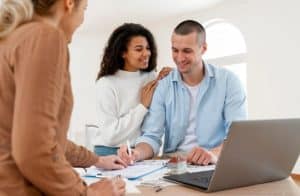 Casal jovem conversa com uma consultora financeira em um ambiente iluminado, ao redor de uma mesa com documentos, canetas, um notebook e uma pequena casa em miniatura. Ambos demonstram atenção e satisfação com as orientações recebidas, sugerindo um momento de planejamento financeiro. A cena representa o interesse em segurança futura e decisões conscientes sobre investimentos, como a contratação de uma previdência privada para garantir estabilidade e tranquilidade no longo prazo.