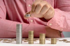 Pessoa com camisa rosa empilhando moedas sobre uma mesa branca, formando colunas de diferentes alturas, simbolizando o crescimento e a organização das finanças. A mão em destaque segura mais moedas, indicando continuidade no processo de economia e planejamento. A imagem transmite a ideia de controle financeiro e estratégias para melhorar a renda mensal de forma estruturada e progressiva.