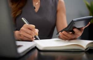 Mulher faz anotações em um caderno enquanto segura uma calculadora, representando o controle do orçamento e a organização das contas como práticas essenciais para manter a saúde financeira em dia.