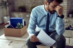 Homem sentado com expressão de preocupação, segurando uma folha de papel e cercado por caixa de pertences profissionais, representando o impacto emocional e financeiro causado pelo aumento da taxa de desemprego.