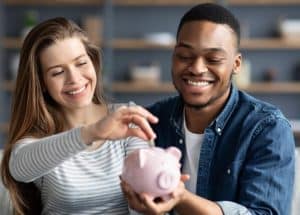 Casal sorridente sentado lado a lado em um ambiente doméstico, demonstrando união e planejamento. A mulher deposita uma moeda em um cofre em formato de porquinho que está sendo segurado pelo homem, simbolizando o ato de economizar juntos. A imagem transmite harmonia, cumplicidade e foco na construção de uma vida financeira saudável e equilibrada.