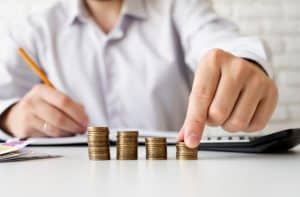 Pessoa organiza moedas empilhadas sobre a mesa enquanto faz anotações com lápis e usa uma calculadora, representando o controle da vida financeira por meio de planejamento e gestão do dinheiro.